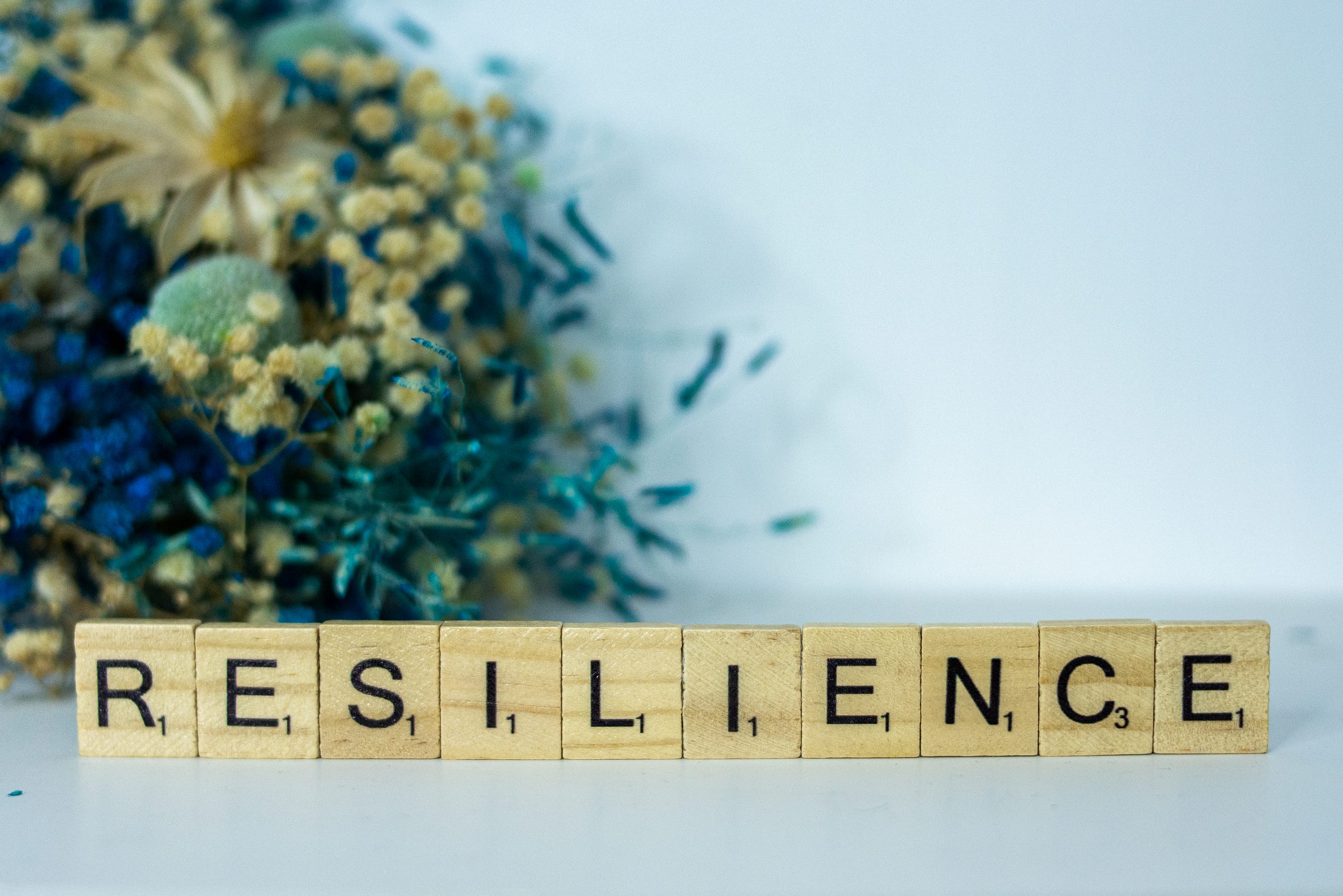 Scrabble tiles spelling out the word RESILIENCE in front of a blurred bouquet of dried flowers